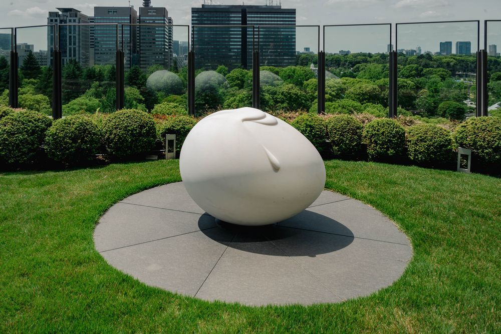 A large smooth white egg-shaped sculpture with subtle organic folds sits on a circular stone plaza in the Park View Garden on the 6th floor of Hibiya Midtown, Tokyo, surrounded by manicured hedges and green lawn, with a dense skyline of high-rise buildings visible beyond a glass railing under a hazy sky.