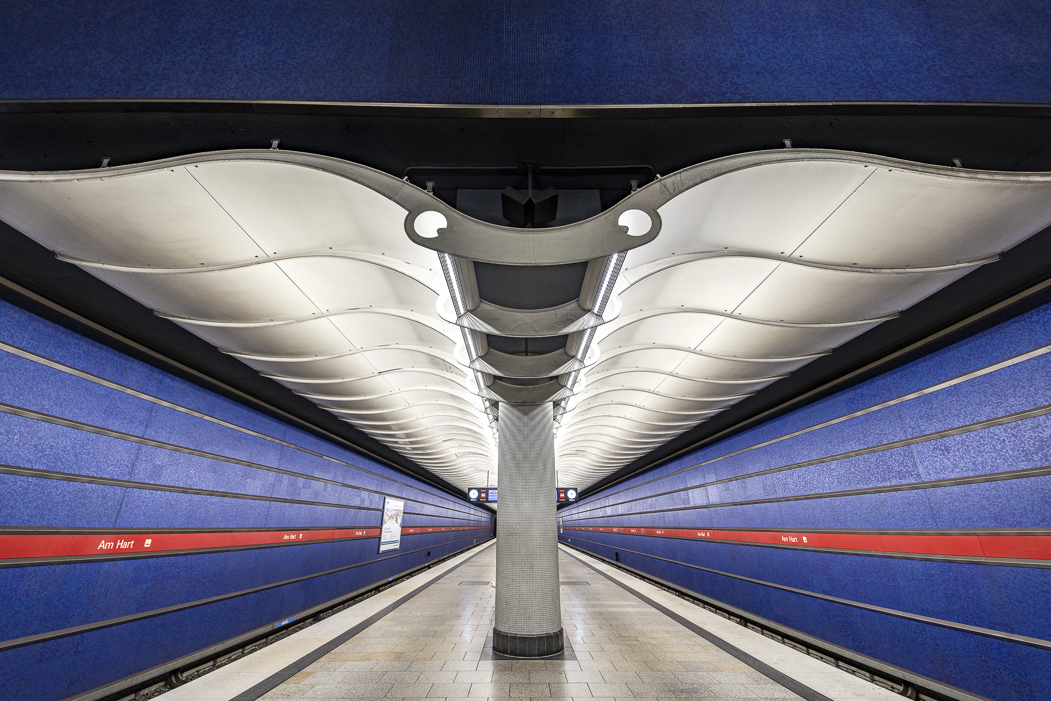 Symetrical wide-angle view of a white canopy on the ceiling at Am Hart U-Bahn station in München, that extends and ondulates on each side of the picture like the wings of a white Manta Ray facing the viewer. Two lit circles in the center look like a pair of eyes. The station is empty and its wall are dark blue, emphasising the impression that a Manta Ray is swimming in the sea.