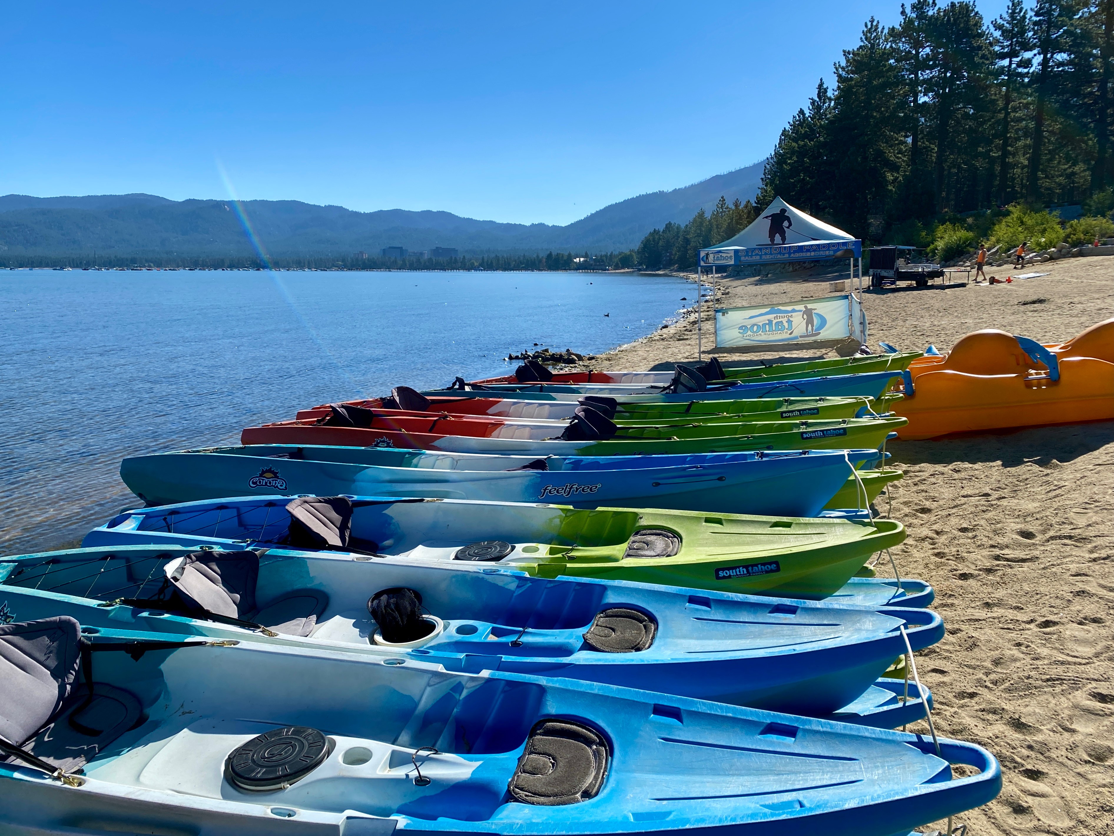 Colorful kayaks for rent are docked on the beach of Lake Tahoe, California. It's early in the day, so the kiosk isn't open yet. Beyond the kayaks are more beach, pine trees, then, of course, Lake Tahoe in the midst of the Sierra Nevada Mountains. The sky is clear and bright blue, as is the lake. South Lake Tahoe