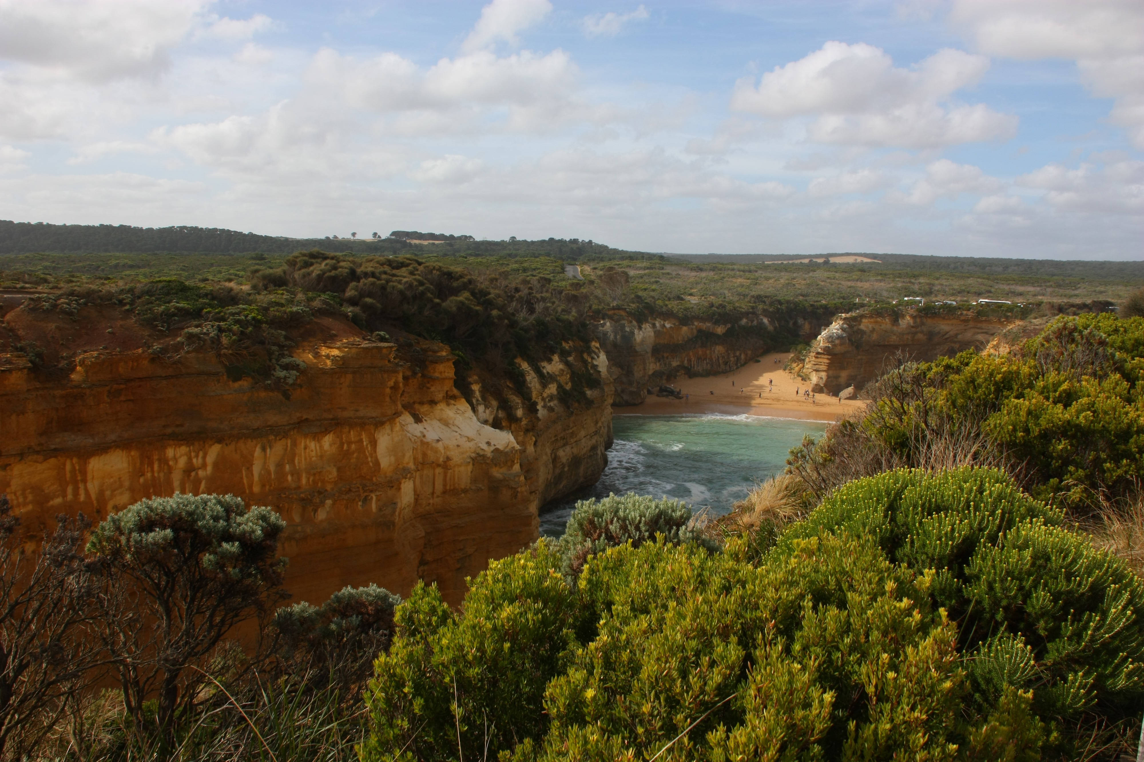 The perspective is from a higher ground, looking out at the flat land of Australia. However, there are high cliffs lining an ocean inlet that culminates in a sandy beach spotted with people that look like specks. The tops of the cliffs are covered with dark green vegetation, the cliffs are red rock and soil, while the inlet/cove water is a soft foam green. The sky is a light blue with white clouds, rather hazy. The entire image gives a sense of how vast the earth's geography is, and when you see the specks of people, it's humbling how insignificant we are.