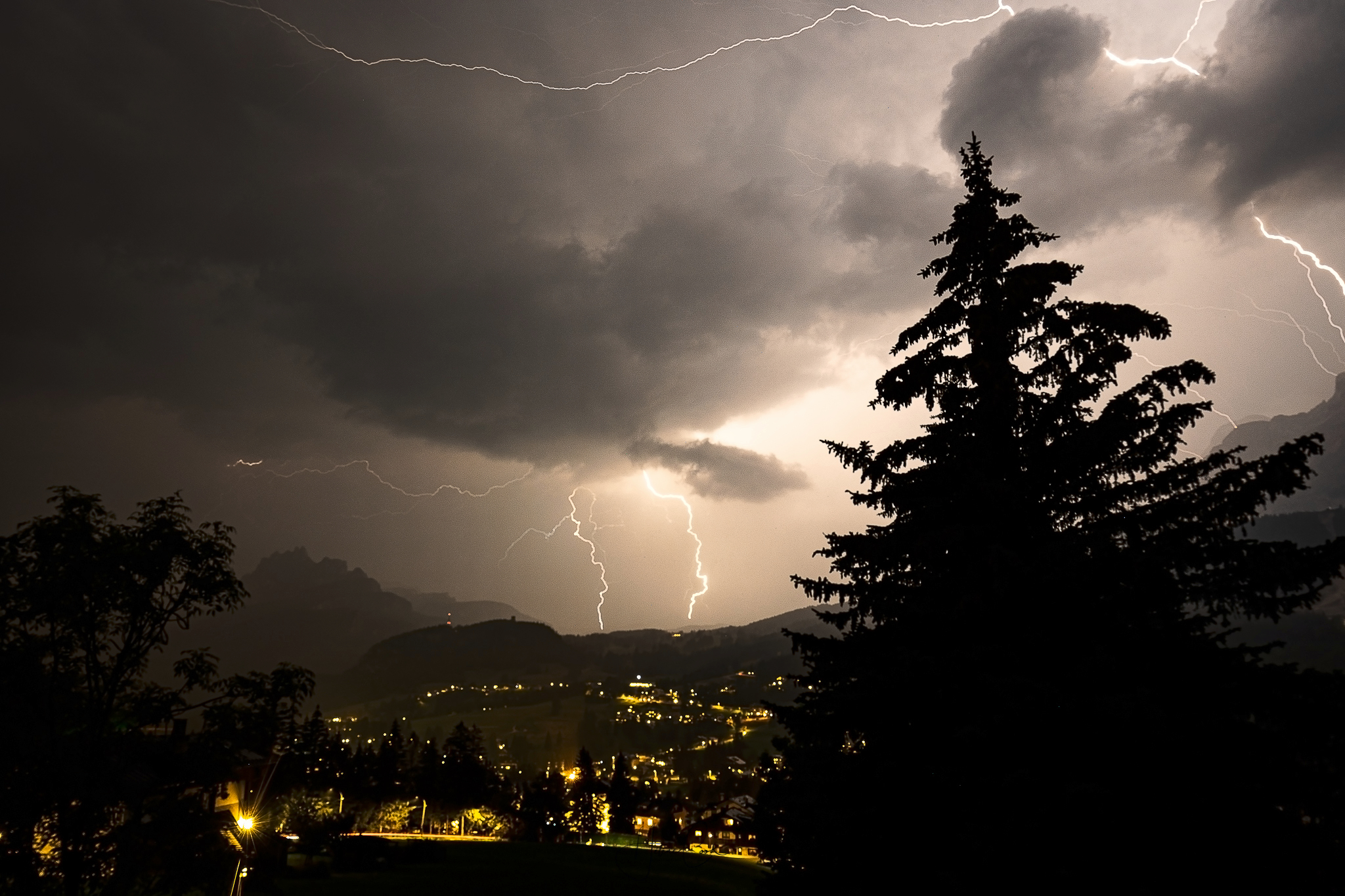 A mountain village under storm in the evening. Wooden houses are lit in the distance, thunder lightnings illuminate the dark sky and reveal dark pine tree shapes in the foregrounds and mountains in the background.