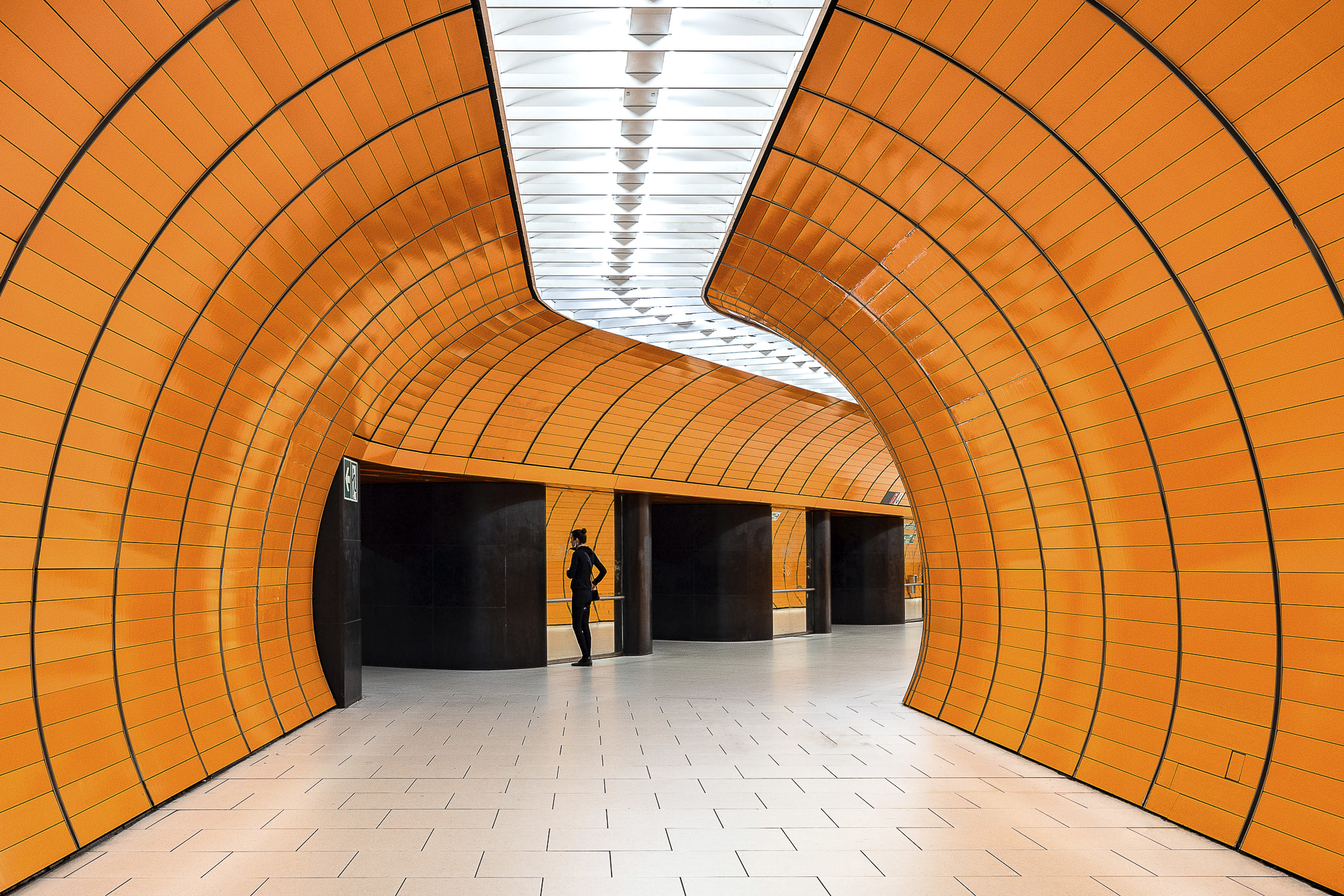 A woman dressed in black watches herself in the mirror in a bright orange curvy tunnel at Munich's Marienplatz metro station. The set of this station conveys vibes from the seventies.
