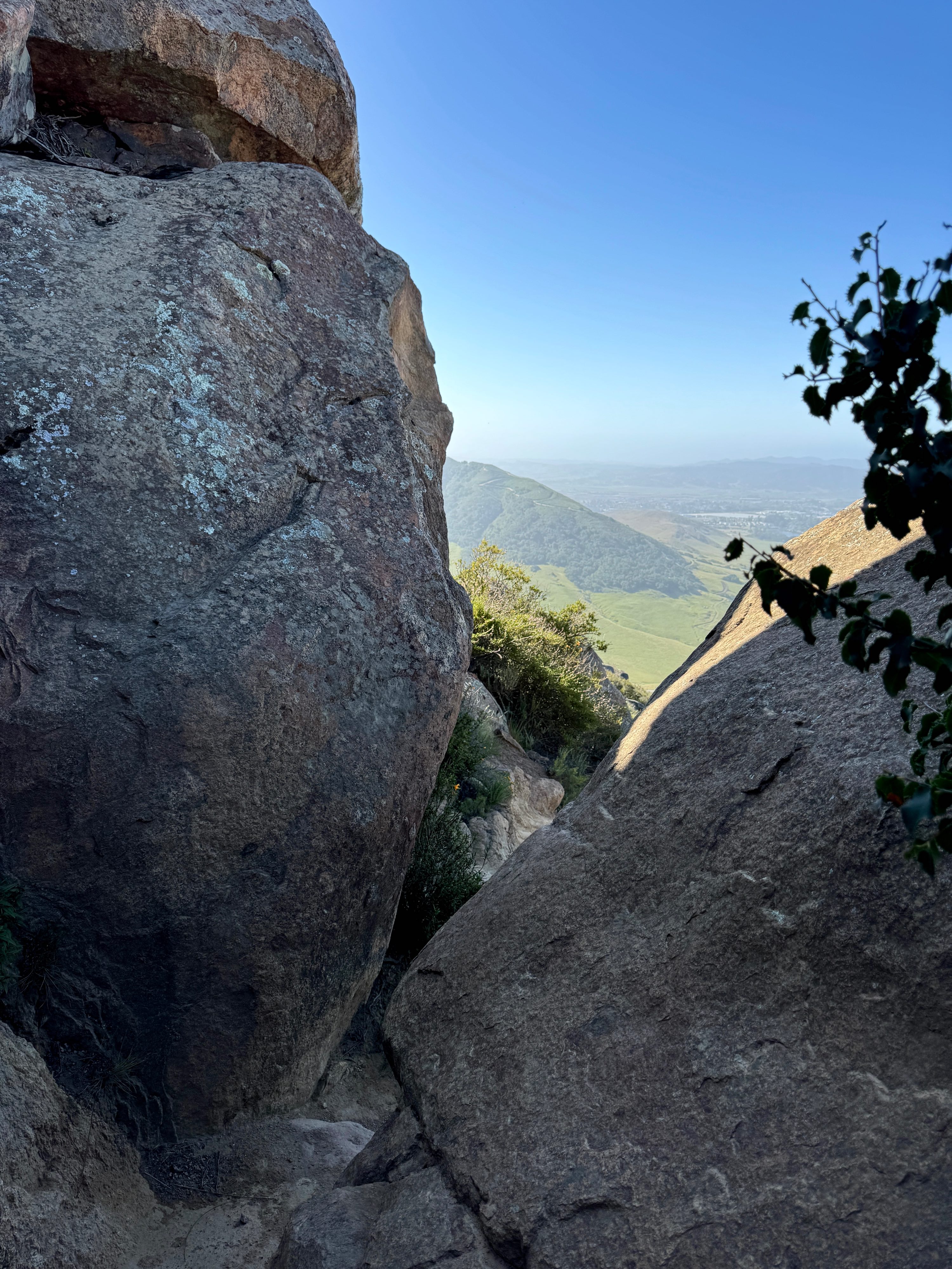 Two huge boulders occupy most of the image, one vertically the entire left side of the photo. There is another smaller boulder that uses just over half of the right side of the image. A hiking trail squeezes between these rocks, though in real life, it's not quite as tight as it looks in the image. Beyond the boulders are some green shrubs, and past those is the view of the green Los Osos Valley and half of Cerro San Luis mountain in San Luis Obispo, California. More mountains are in the hazy distance, and the sky is blue once you get above the haze. Small silhouetted shrubbery is in the center right edge foreground, overlapping the smaller boulder. Both boulders are in the shadow of Bishop Peak, the tallest of the Nine Sisters.
