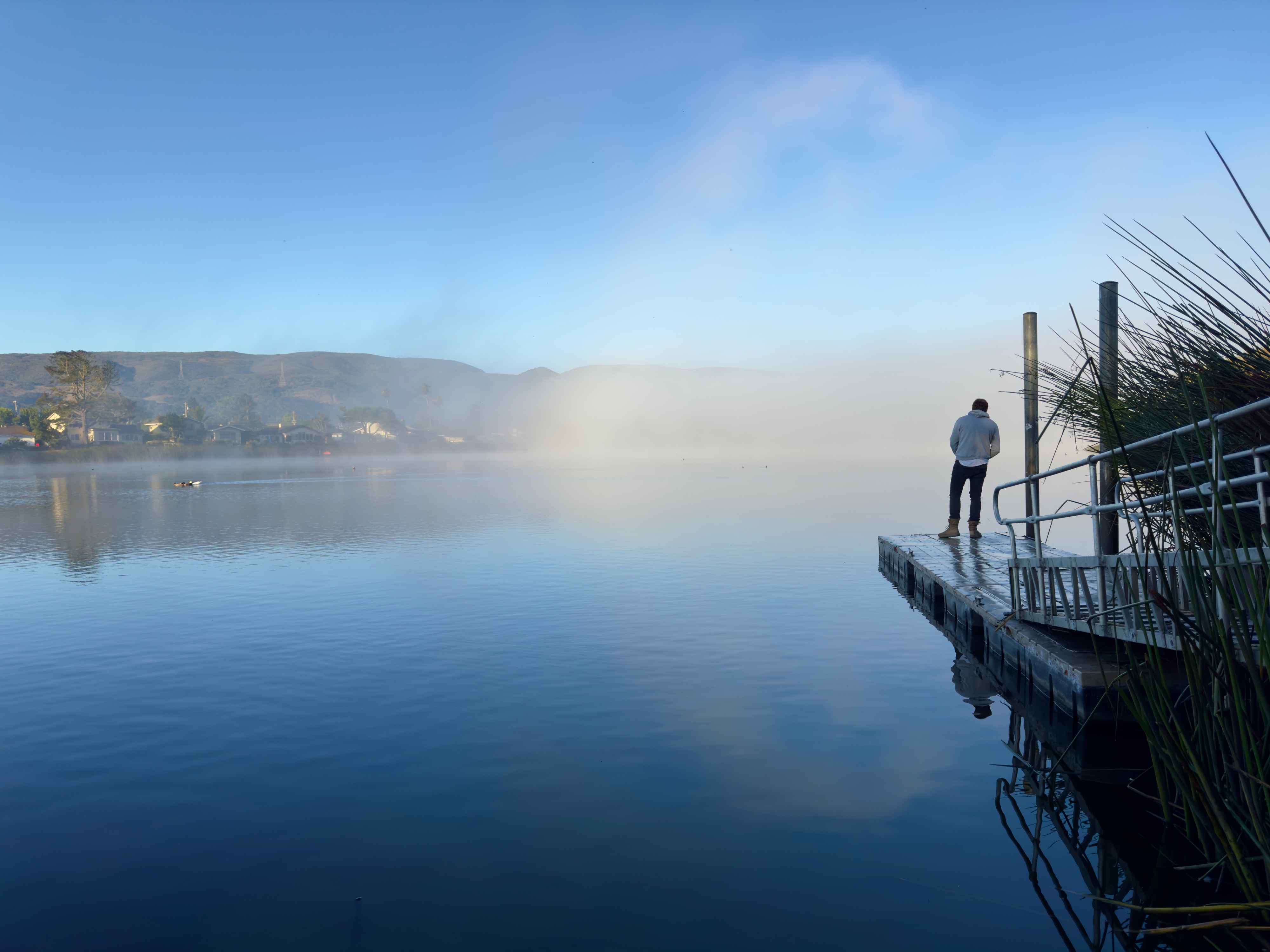 A silhouetted man is standing at the end of a small dock fishing in Laguna Lake. It's early in the morning with light fog which has formed a partial fogbow over the lake. The sky and lake are both a brilliant blue. Mountains and houses are across the lake and beyond. San Luis Obispo, California