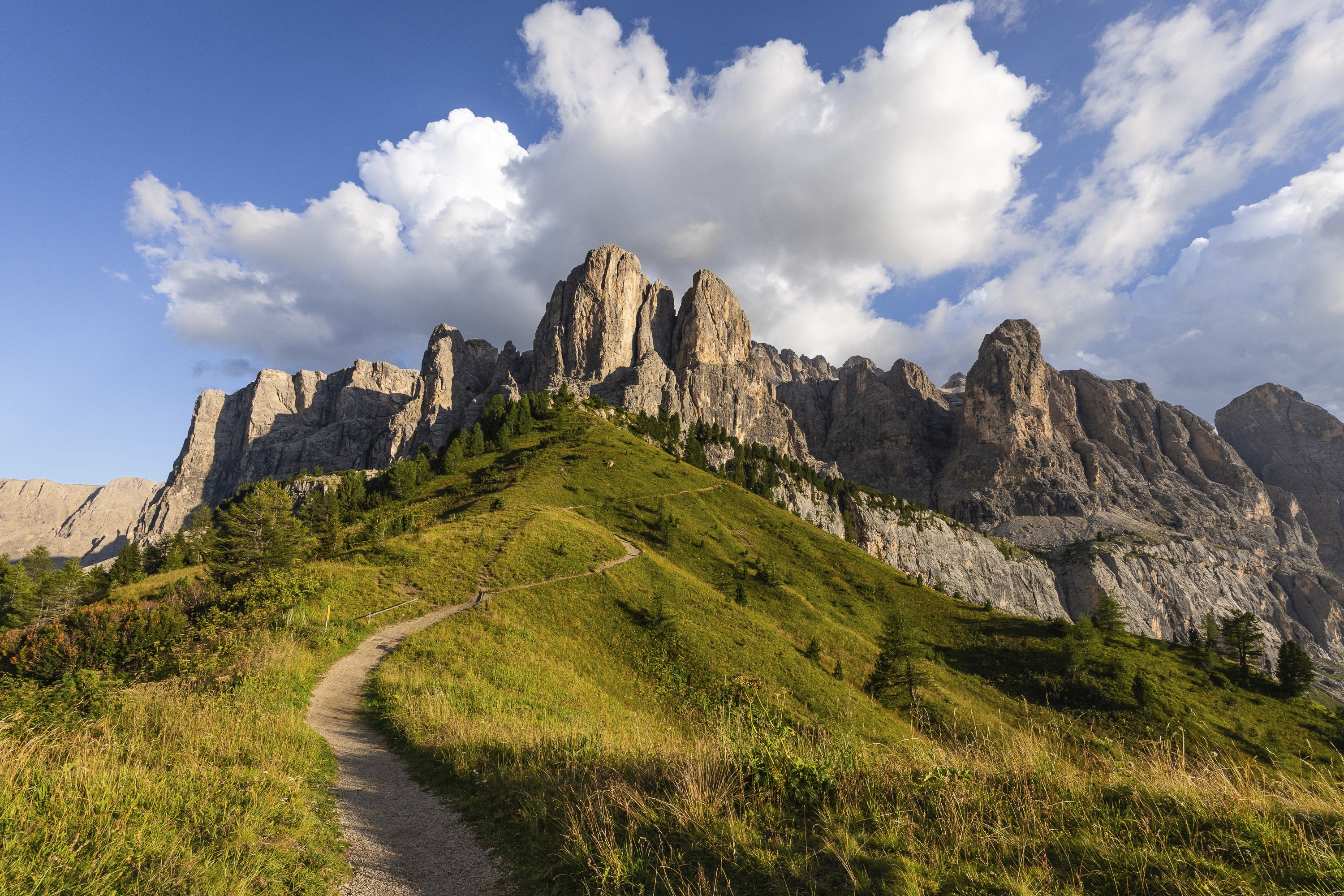 Val Gardena Dolomites mountain picks at golden hour before sunset in summer. A sinuous path leads to majectic picky rocks in hilly fields under a beautiful bluesky with a big white sky ornating the picks like a crown.