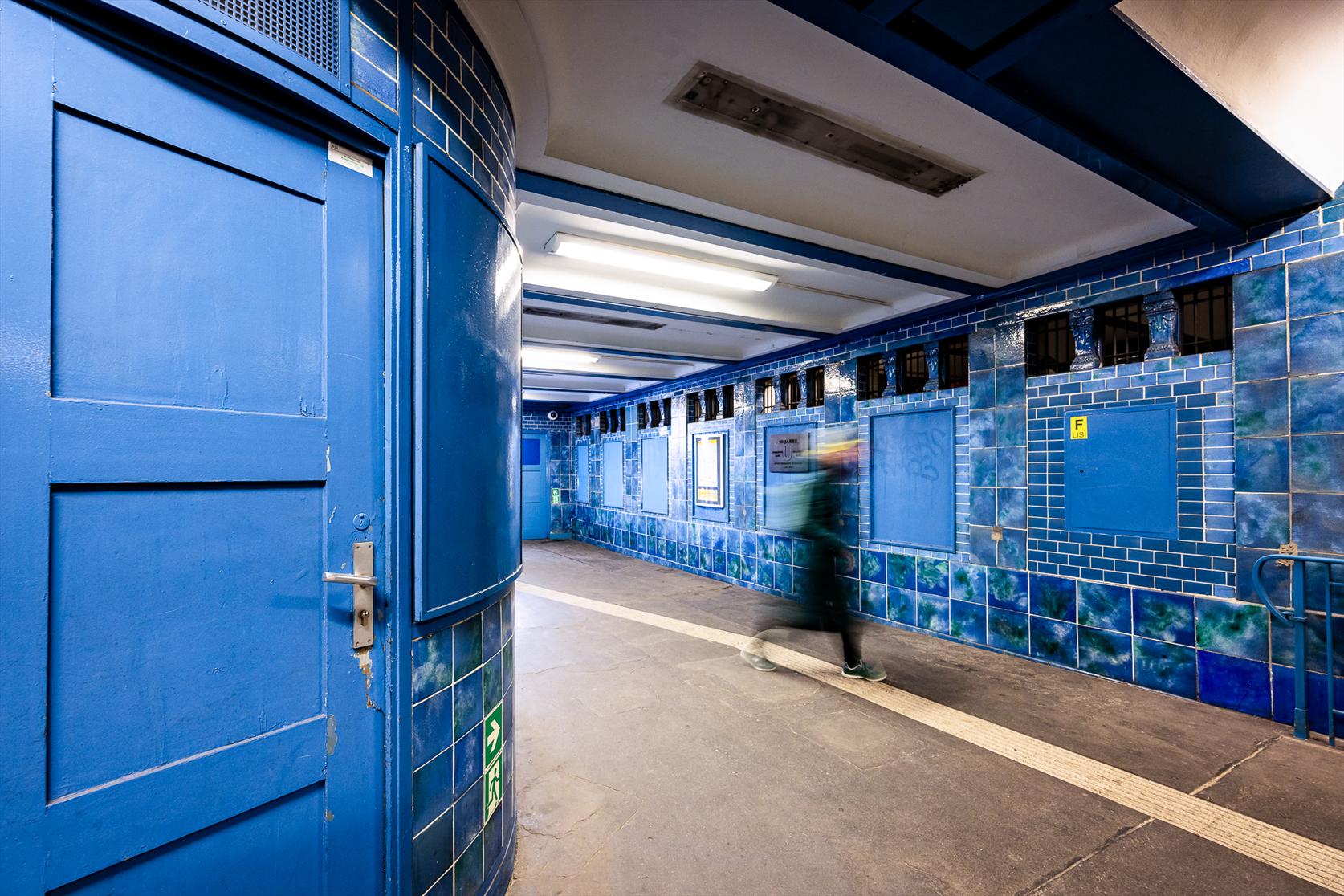 A woman dressed  in dark clothes is hurrying through a corridor decorated with blue tiles and blue wooden doors at Bayerischer Platz metro station in Berlin. Her silhouette is blurry due to the use of a slow shutter speed that enhanced her movement against the stady background.