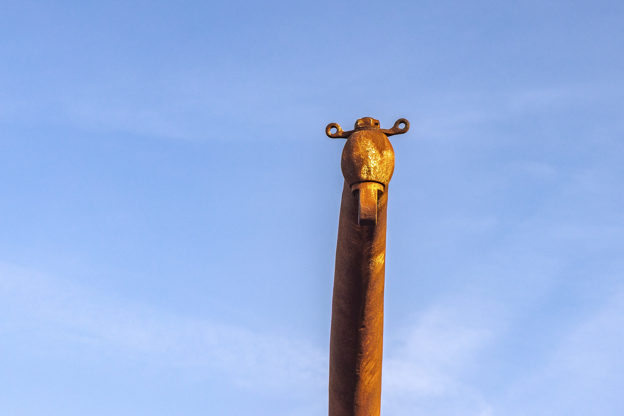 The top of a rusty orange metal crane with round features and little loops on the top which ressembles a giraffe, as seen from below with the blue sky in the background.
