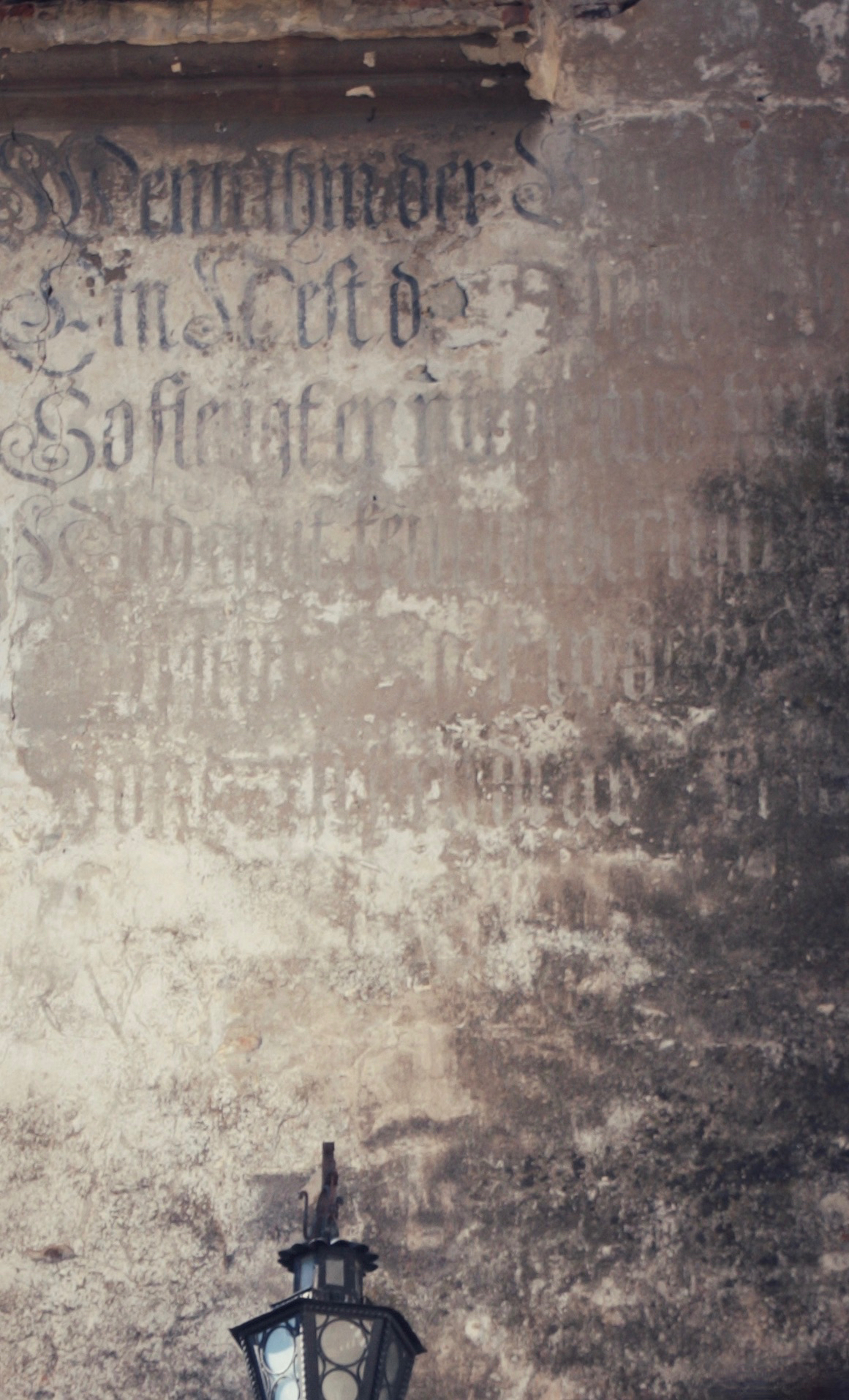 Cropped portion of an exterior castle wall that has faded and chipped paint plus dark stains, possibly mold. There is some old Latin script on the wall that is heavily damaged. Its color ranges from black to so faded that it's light colored.  The wall is colors of beige, taupe, dark brown and black. Just left of center at the bottom of the image is a cropped lantern in black metal and clear glass. Hint: this castle is in Romania.