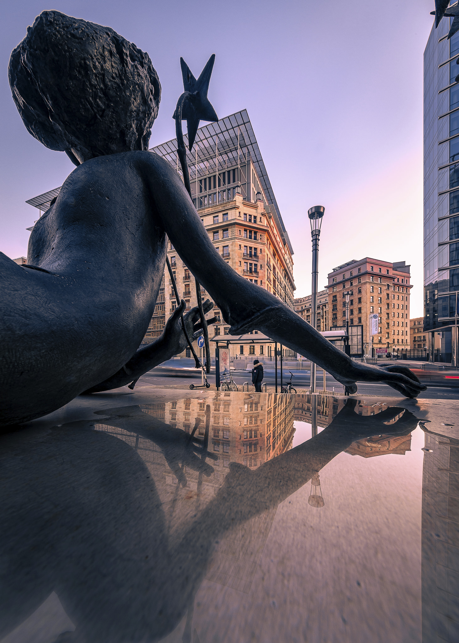 A statue of a woman lying on her belly and resting on her forearms holding a stick with a star at the top, on Rue de la Loi in Brussels. The statue seems to be looking at the direction of the Residence Palace building which is recognisable in the background and lit by the sunset. The statue is part of an art piece by sculptor René Julien.