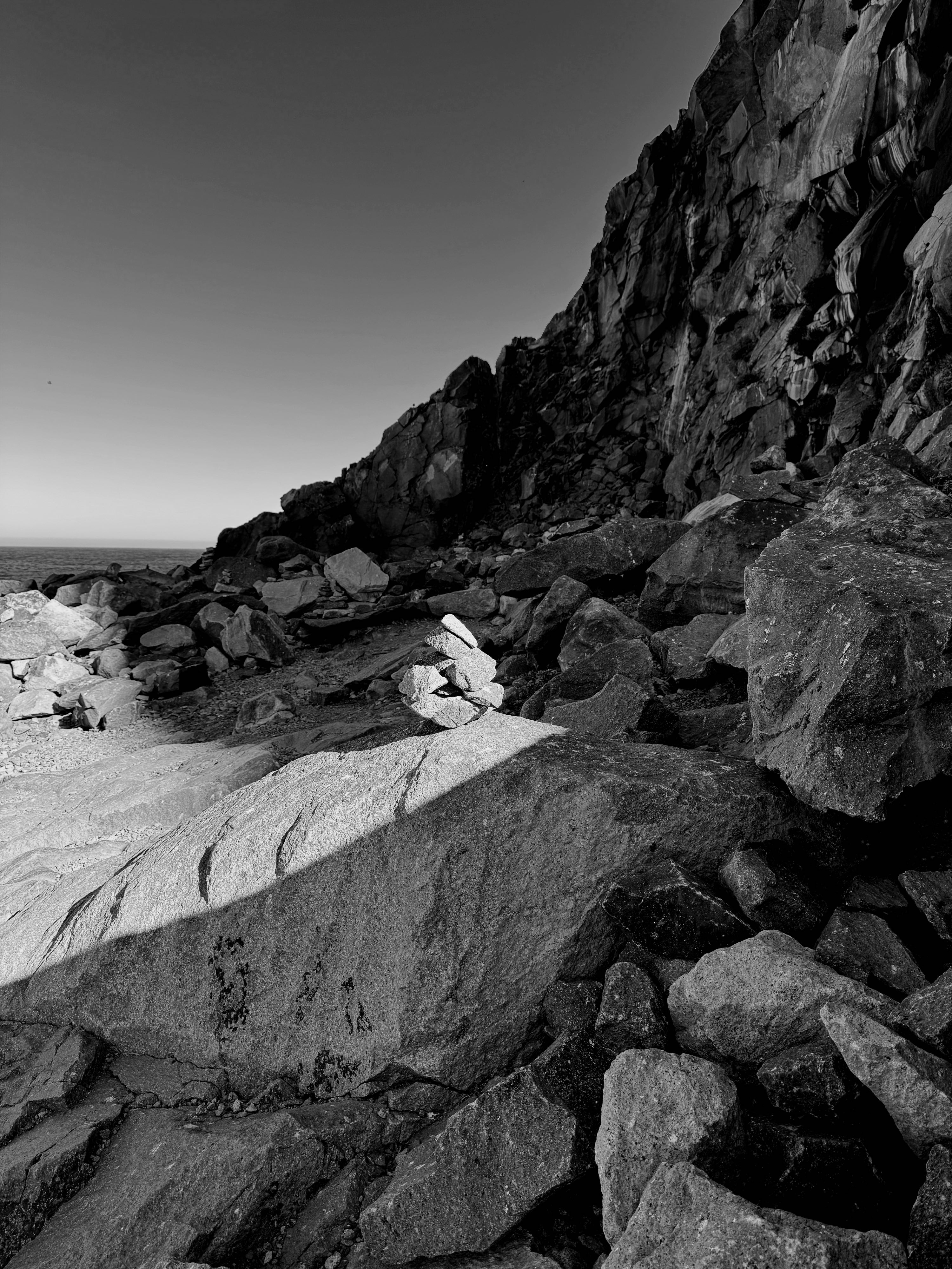 A stack of smaller rocks (a cairn) sits on top of a bigger rock that is at the base of a really big rock...Morro Rock. Morro Rock is in its own shade as are most of the rocks at the base, but the sun is highlighting the cairn, creating quite a contrast with Morro Rock. A sliver of the ocean is behind Morro Rock, and the sky is clear.
