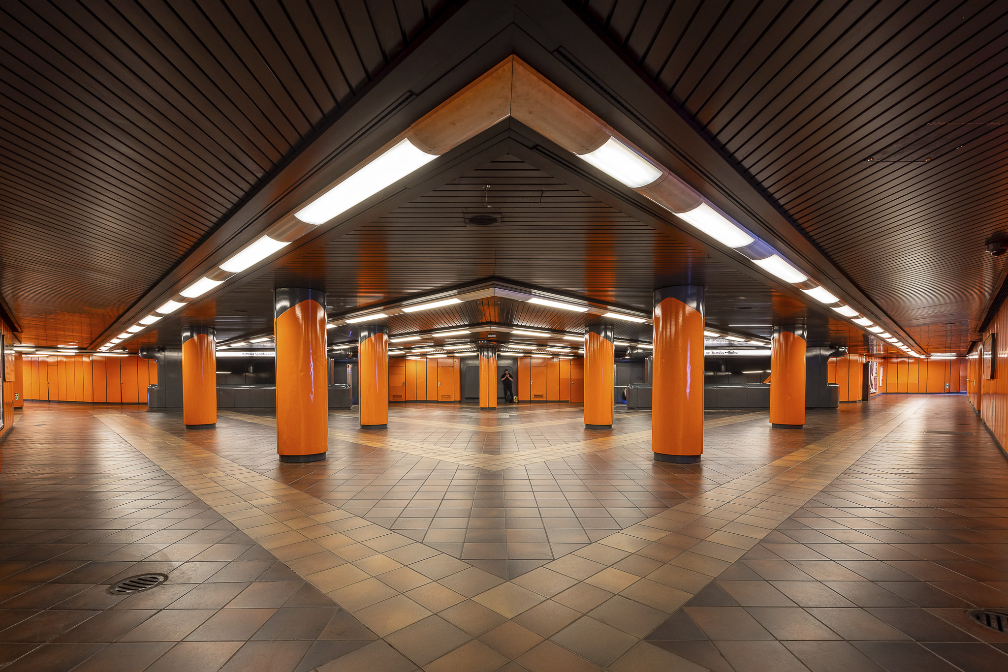Wide angle view of upper floor at Siemmensdamm metro station in Berlin, featuring massive orange pillars and white neon lights on the ceiling arranged in a series of V-shapes. A man in black cloths waiting in a corner against a black wall is almost invisible.
