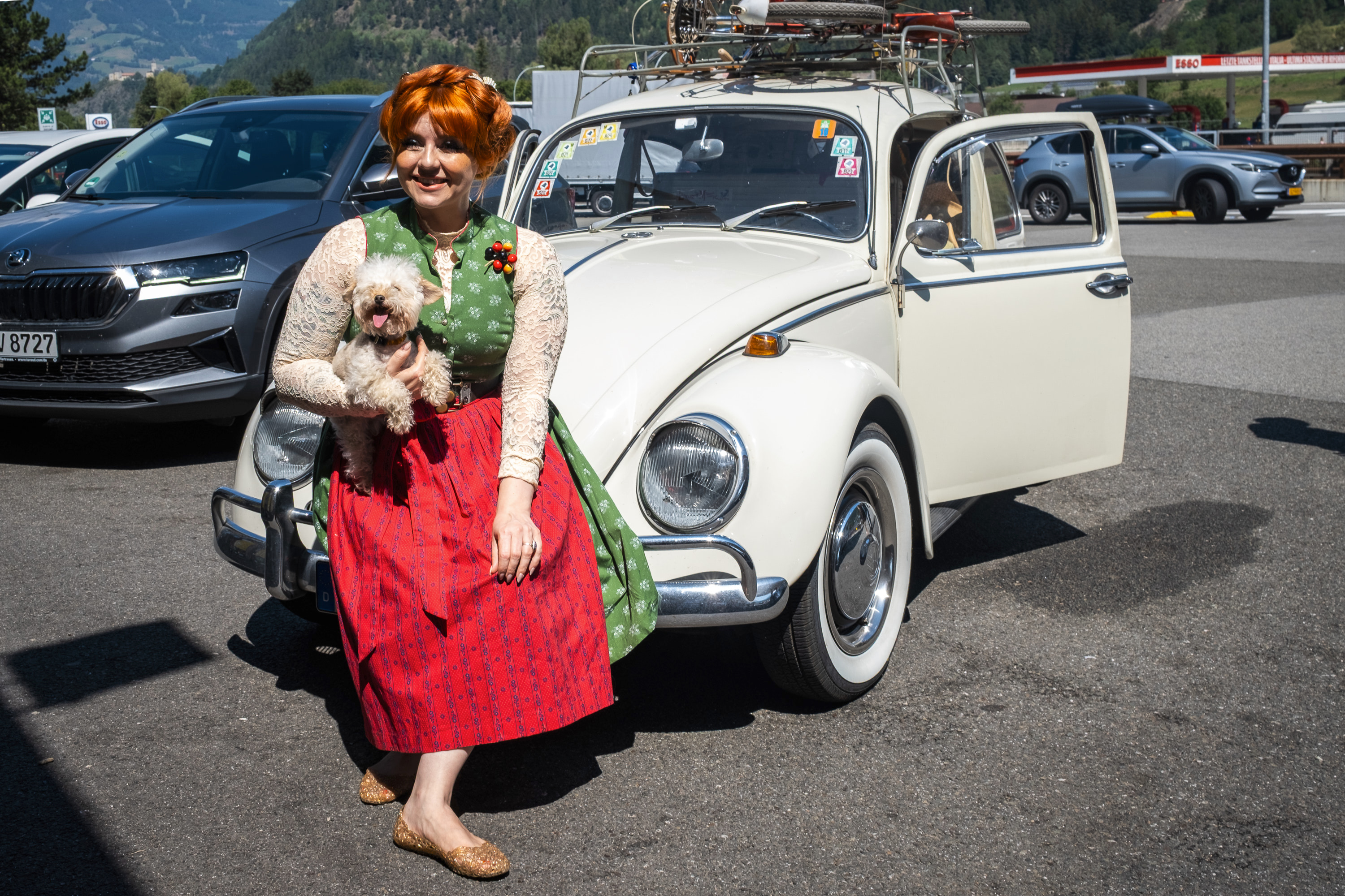 A woman with red braided hair tied up on her head dressed in a green dress with white flowers and a red apron with a cute fluffy doggy on her lap, seated on the hood of a white beatle Volkswagen car, at a motorway station.