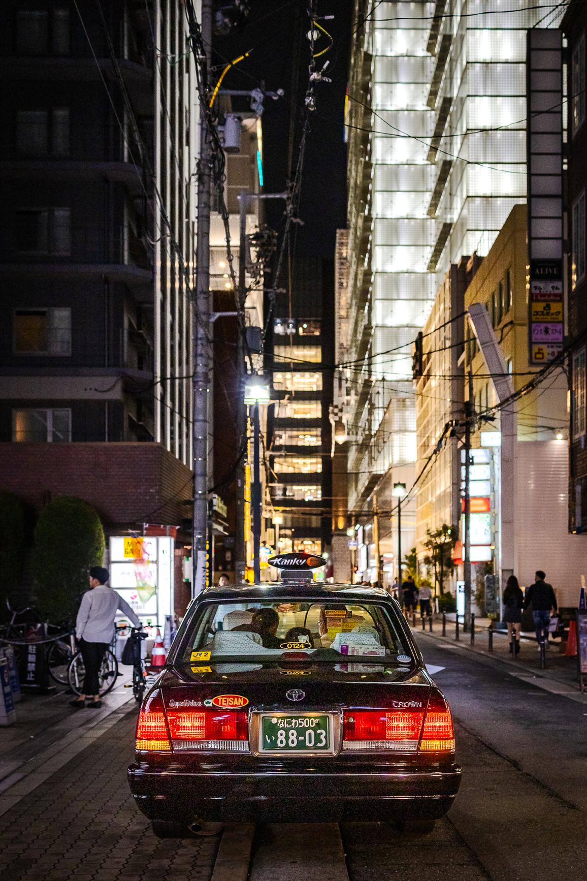 An old black Toyota crown super deluxe taxi, seen from behind, in a narrow street of Osaka surrounded by illuminated highrise buildings, at night.